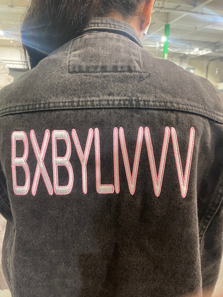BXBYLIVVV Embroidered on a charcoal denim jacket in White text outlined in pink satin stitch