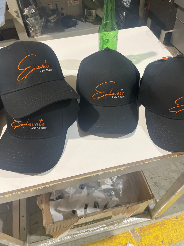 4 Hats on a table with orange embroidered text "Elevate"  and white text "law group" underneath 